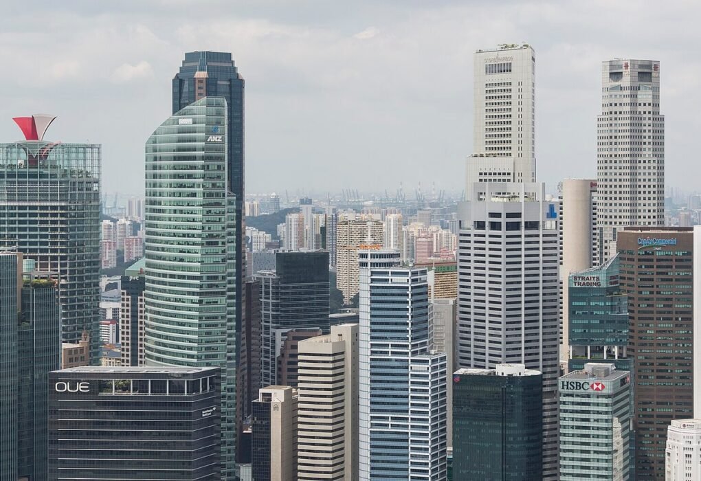 Singapore's Central Business District skyline at Marina Bay, featuring a dense cluster of high-rise commercial and financial towers including buildings bearing the ANZ, HSBC, OUE, NTUC, Straits Trading and Maybank signs, viewed from the Marina Bay Sands Hotel observation deck under a partly cloudy s