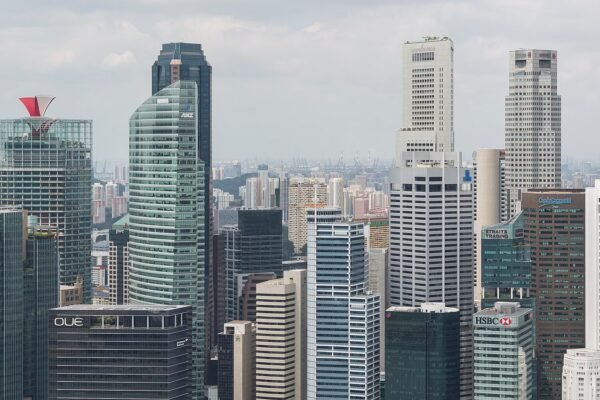 Singapore's Central Business District skyline at Marina Bay, featuring a dense cluster of high-rise commercial and financial towers including buildings bearing the ANZ, HSBC, OUE, NTUC, Straits Trading and Maybank signs, viewed from the Marina Bay Sands Hotel observation deck under a partly cloudy s