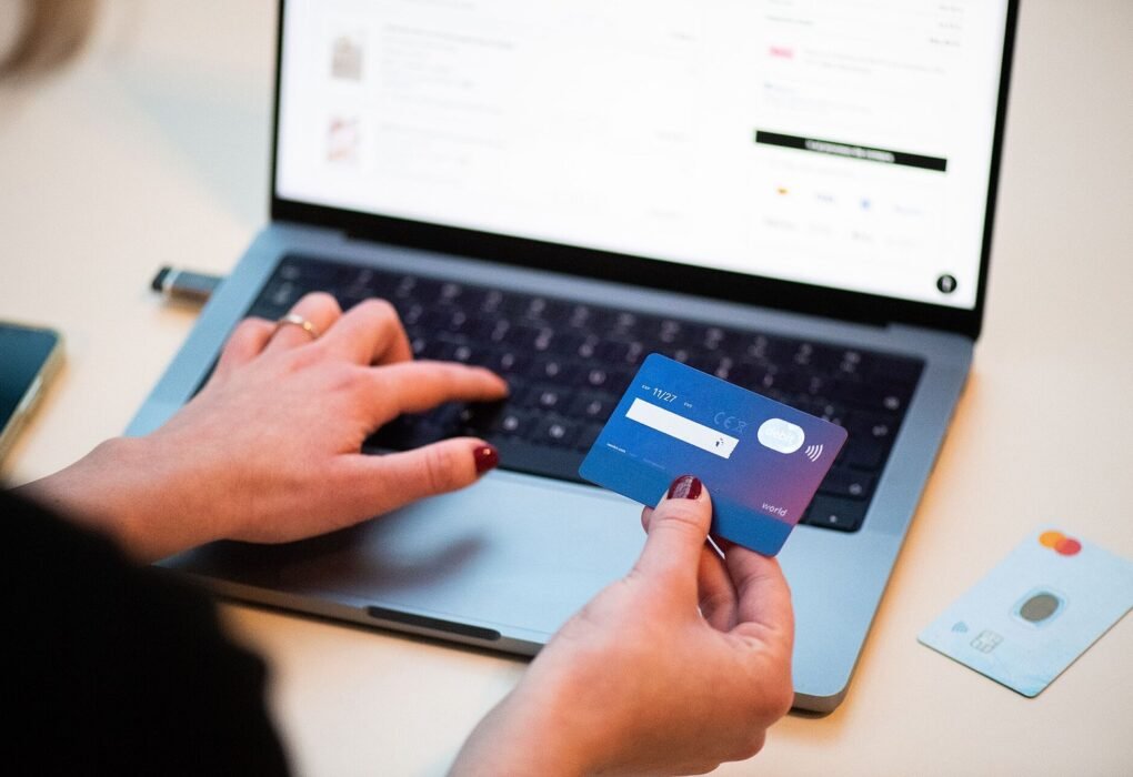 A person holding a bank card while typing on a laptop, with an e-commerce website visible on the screen and a second card resting on the desk nearby