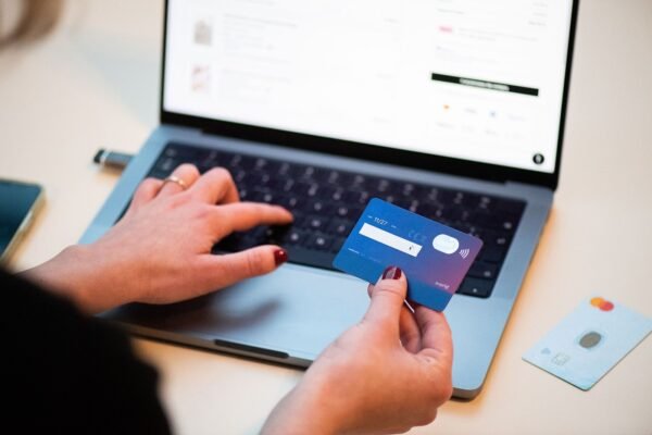 A person holding a bank card while typing on a laptop, with an e-commerce website visible on the screen and a second card resting on the desk nearby