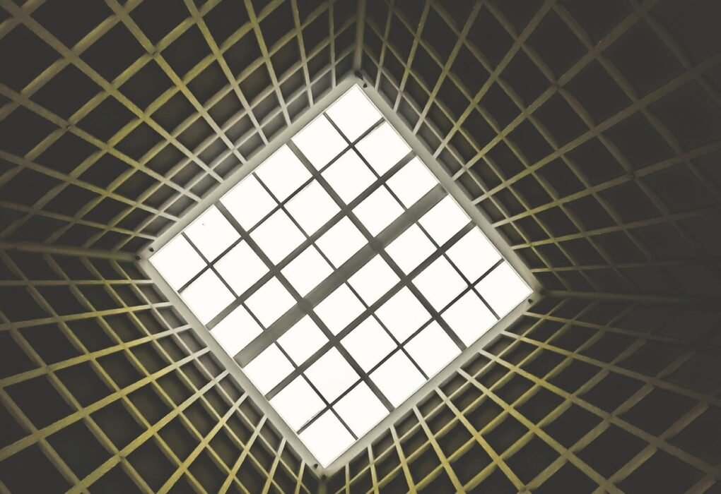 Upward-looking view of a geometric architectural ceiling featuring a grid-patterned skylight at the centre, surrounded by converging structural ribs radiating outward in a dark interior space
