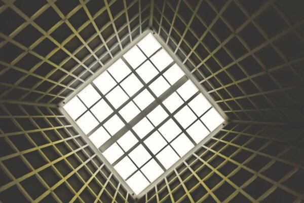 Upward-looking view of a geometric architectural ceiling featuring a grid-patterned skylight at the centre, surrounded by converging structural ribs radiating outward in a dark interior space