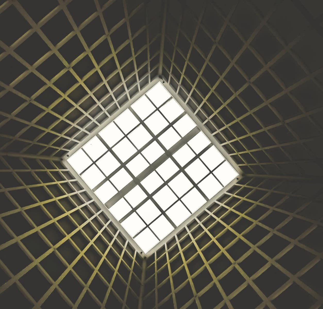 Upward-looking view of a geometric architectural ceiling featuring a grid-patterned skylight at the centre, surrounded by converging structural ribs radiating outward in a dark interior space