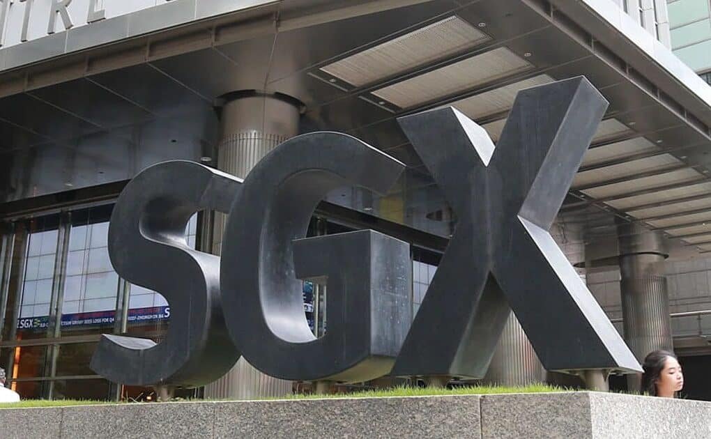 Large black SGX letters forming a sign outside the Singapore Exchange building, with people walking nearby and the building's glass facade visible in the background