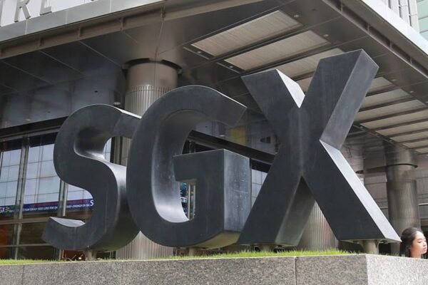 Large black SGX letters forming a sign outside the Singapore Exchange building, with people walking nearby and the building's glass facade visible in the background